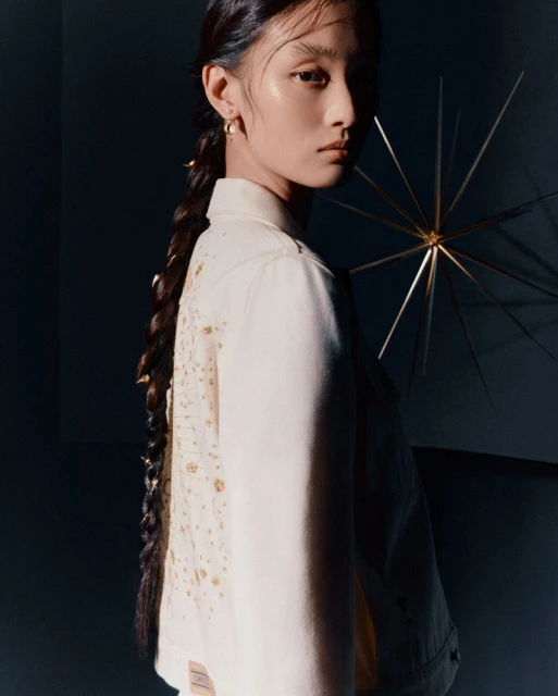 Portrait of a young woman with a long braid wearing a decorated off-white denim jacket against a dark studio backdrop.