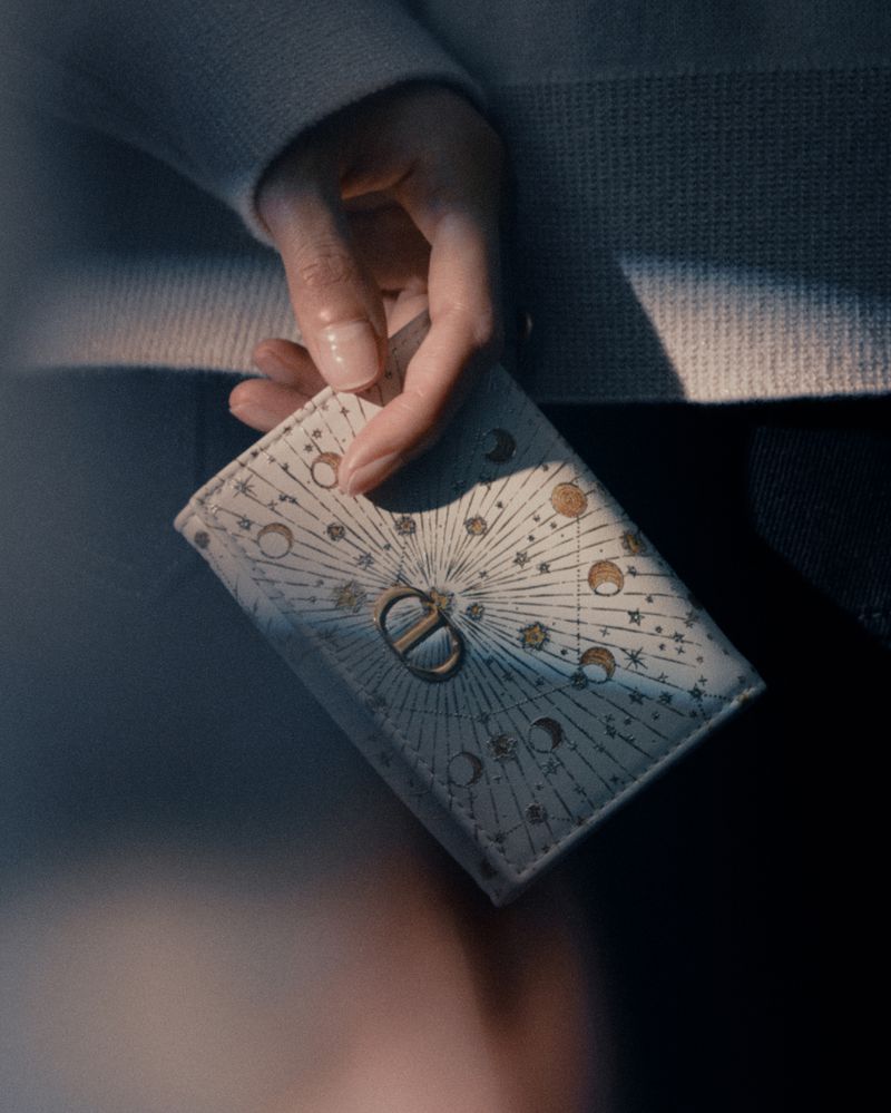 Woman holding a Dior wallet. The cream-colored wallet features a pattern of celestial bodies and the Dior logo in the center.