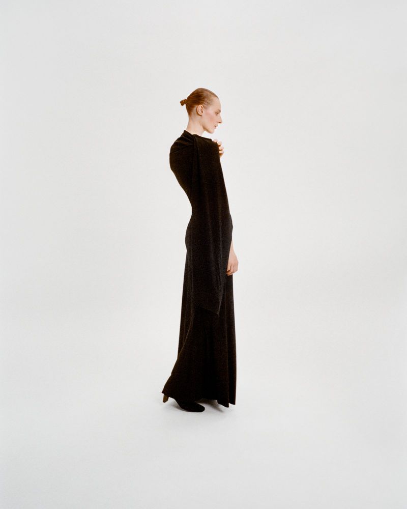 A slender woman in a black floor-length dress poses in a minimalistic studio with a plain white backdrop, showcasing fashion and elegance.