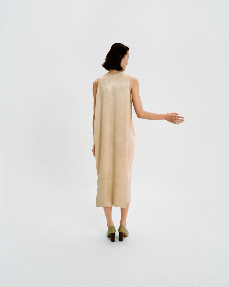 Back view of woman in tan midi dress and green block heel shoes in a minimalist white setting. Fashion shoot, clean style.