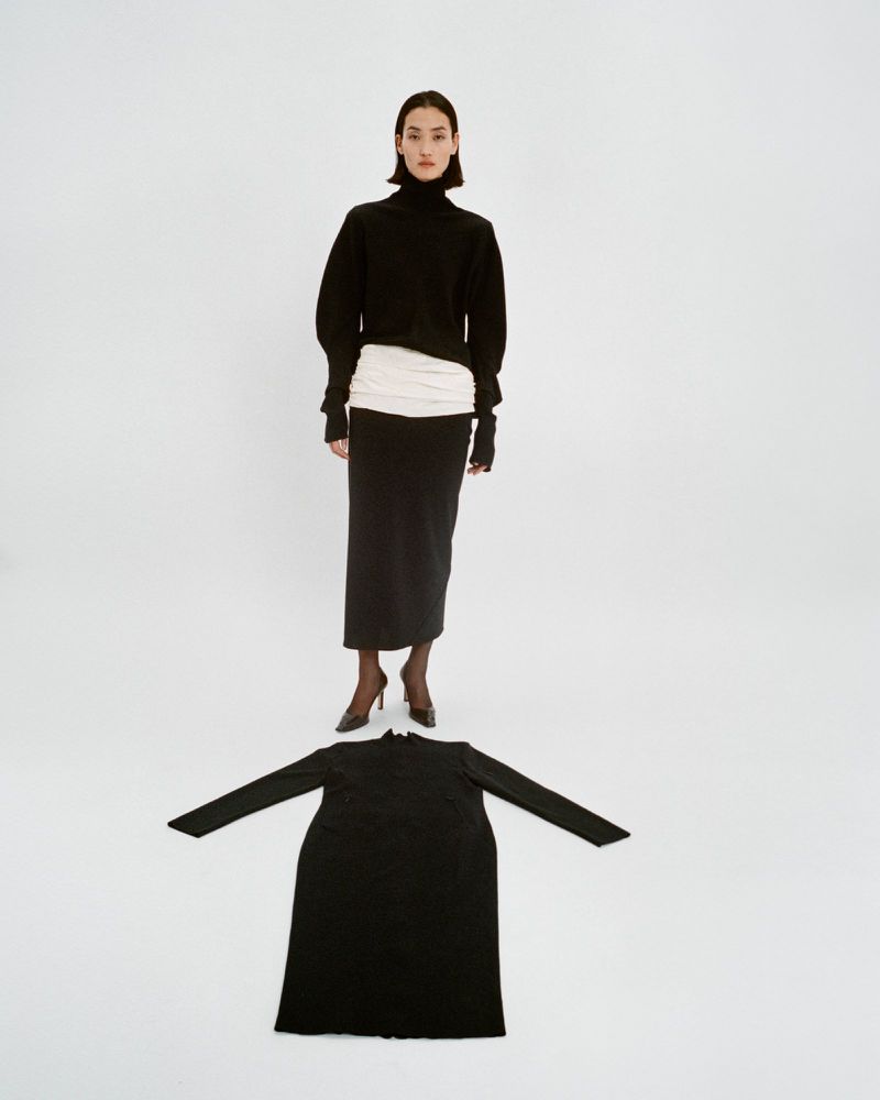 Studio shot of a stylish woman in a black turtleneck and long skirt standing above a black dress on a white seamless backdrop.