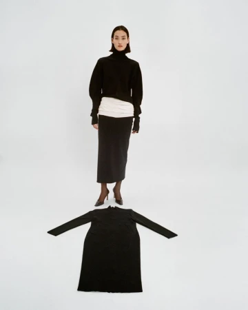 Studio shot of a stylish woman in a black turtleneck and long skirt standing above a black dress on a white seamless backdrop.