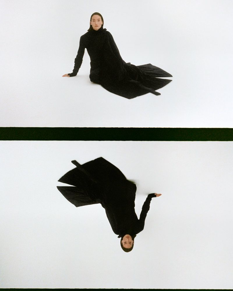 Woman wearing a long black dress sits on a white surface, reflected below by a black stripe, in a studio fashion shot.