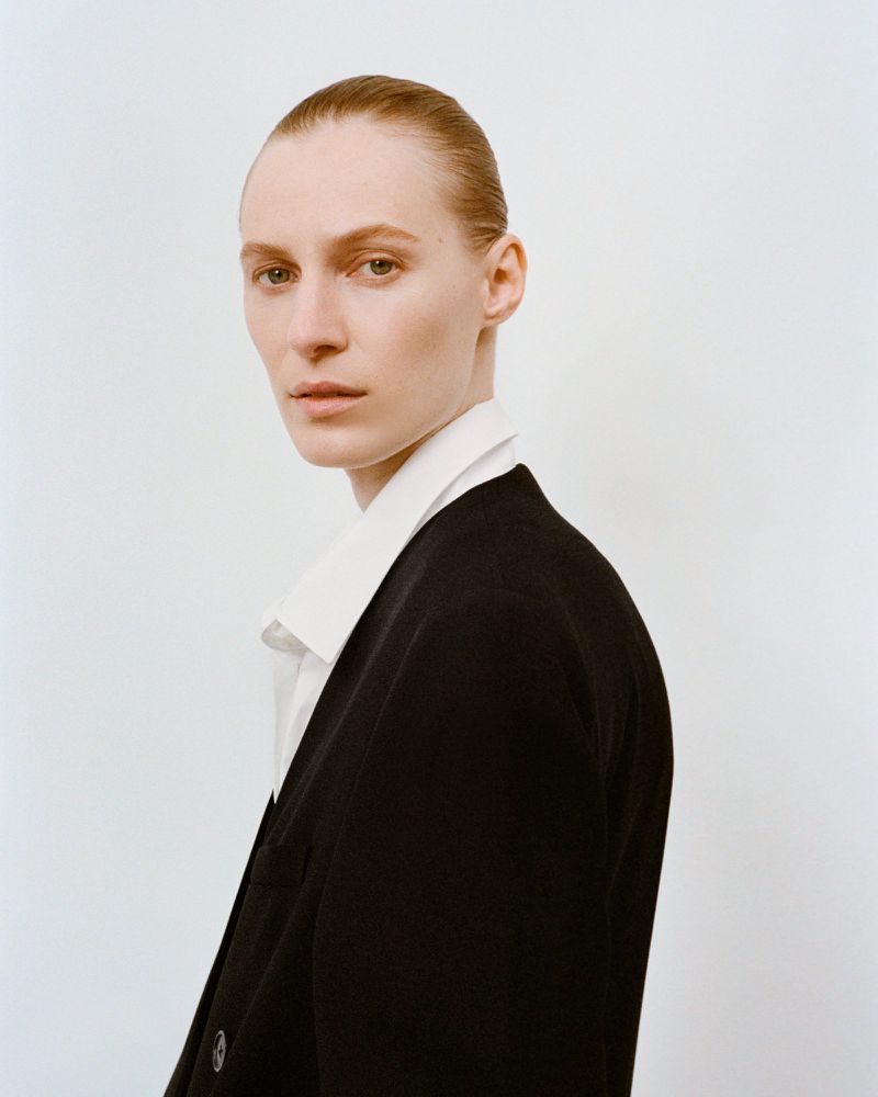 Elegant woman in a black blazer and white collared shirt stands against a white background, looking directly at the camera in a studio portrait.