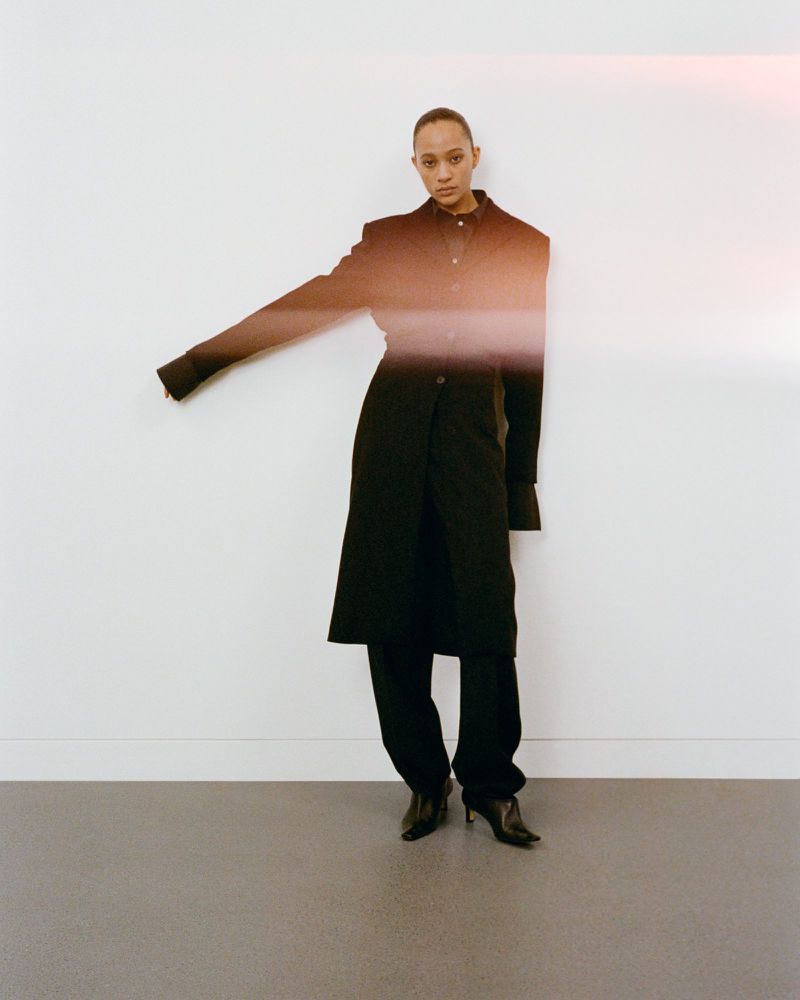 Fashion shot of a slim adult woman in a dark coat and pants against a white wall, with an outstretched arm and neutral expression.