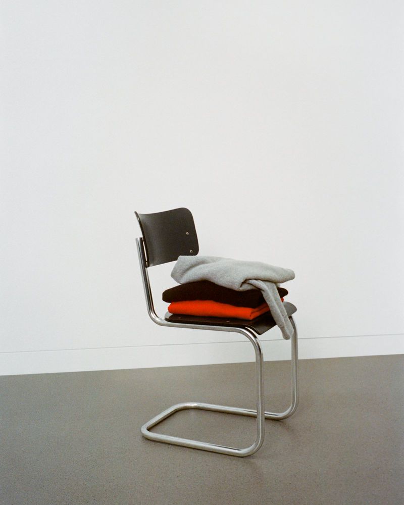 Modern chrome chair with stacked folded knitwear in gray, black, and rust orange against a minimalist white wall and gray floor.