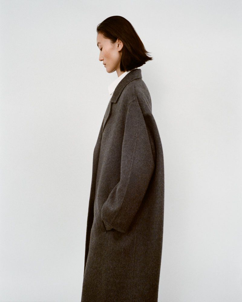 Side profile of a woman with short dark hair wearing a long grey wool coat in a minimalist, neutral-toned indoor setting.