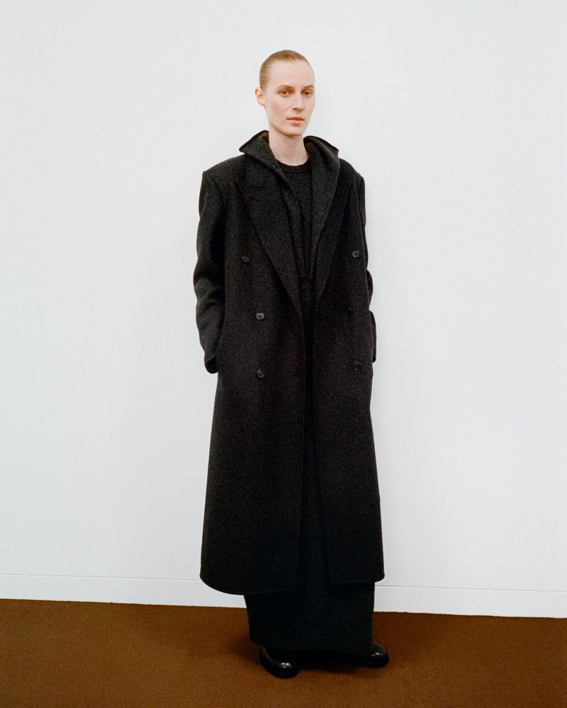 Full-length studio shot of an androgynous person in a long dark gray double-breasted wool coat, dark clothing, standing against white wall.