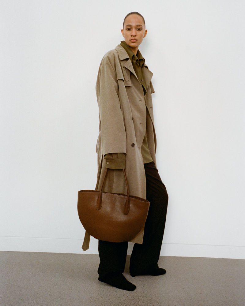 Fashion portrait of a woman wearing a trench coat and carrying a leather tote bag against a white wall, studio lighting.