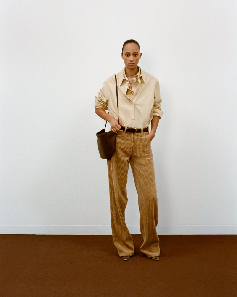 Adult female in a tan shirt and trousers, carrying a dark brown bucket bag, poses indoors against a white and brown background.