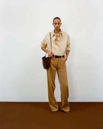 Adult female in a tan shirt and trousers, carrying a dark brown bucket bag, poses indoors against a white and brown background.