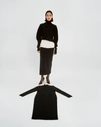Model wearing a black turtleneck and midi skirt standing above a matching black garment laid flat on a white studio background.