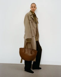 Model in camel trench coat and dark trousers holding a large brown leather tote bag against a white studio background.