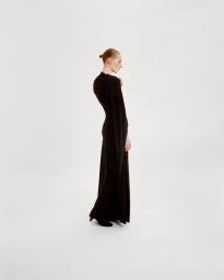 Model standing in profile wearing a long black dress against a plain white studio backdrop.