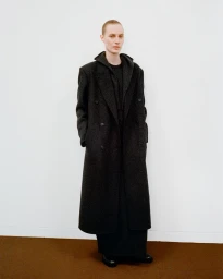 Model standing against a plain white wall wearing a dark wool coat and matching trousers for The Row Fall 2025 collection.