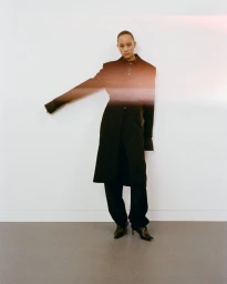 Model standing against a plain white wall wearing a long dark coat and trousers, captured with a soft light leak effect across the frame.