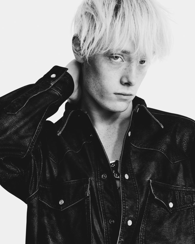 Monochrome portrait of a young adult male with blond hair wearing a dark leather jacket, posing against a white backdrop in a studio.