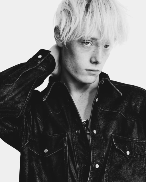 Monochrome portrait of a young adult male with blond hair wearing a dark leather jacket, posing against a white backdrop in a studio.