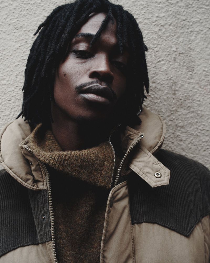 Portrait of Black man with dreadlocks wearing neutral-colored puffer jacket and turtleneck sweater against concrete wall, medium shot.