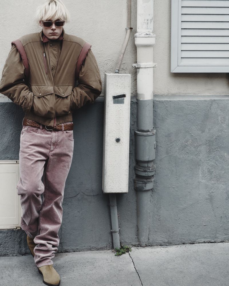 Man poses against a textured wall in jacket and pink pants. He wears sunglasses, and beige boots, in an outdoor urban setting.