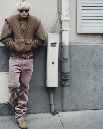 Man poses against a textured wall in jacket and pink pants. He wears sunglasses, and beige boots, in an outdoor urban setting.