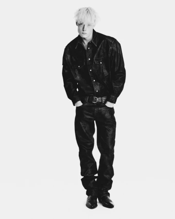 Full-length portrait of a male model with bleached blonde hair wearing a dark leather shirt and matching trousers against a white backdrop.