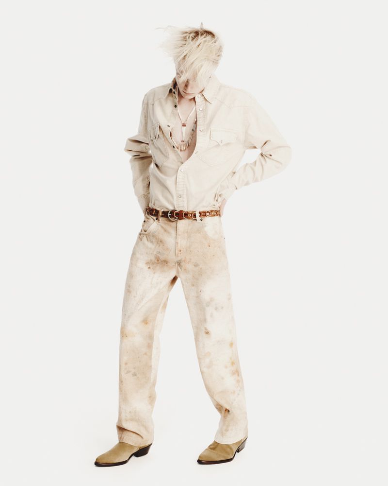 Full-length studio shot of adult male with blonde hair, wearing a tan button-down shirt, brown belt, distressed denim pants, and cowboy boots.