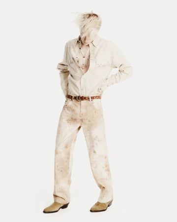 Full-length studio shot of adult male with blonde hair, wearing a tan button-down shirt, brown belt, distressed denim pants, and cowboy boots.