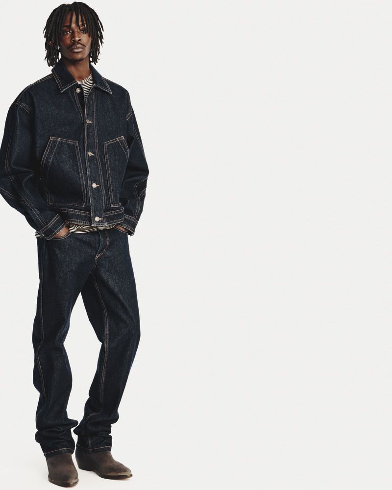 Full-length studio shot of a male fashion model wearing a dark denim jacket and jeans, styled with brown suede boots, against a white backdrop.