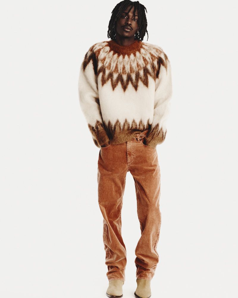 Male model stands in a full shot, wearing a white and brown patterned sweater, tan corduroy pants, and tan boots against a white backdrop.