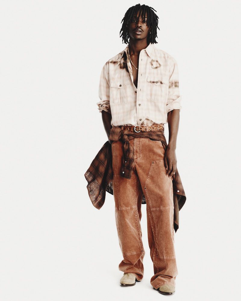 Full shot of a young adult Black male model in a light-colored shirt, brown pants, and brown shoes against a white backdrop in a studio.