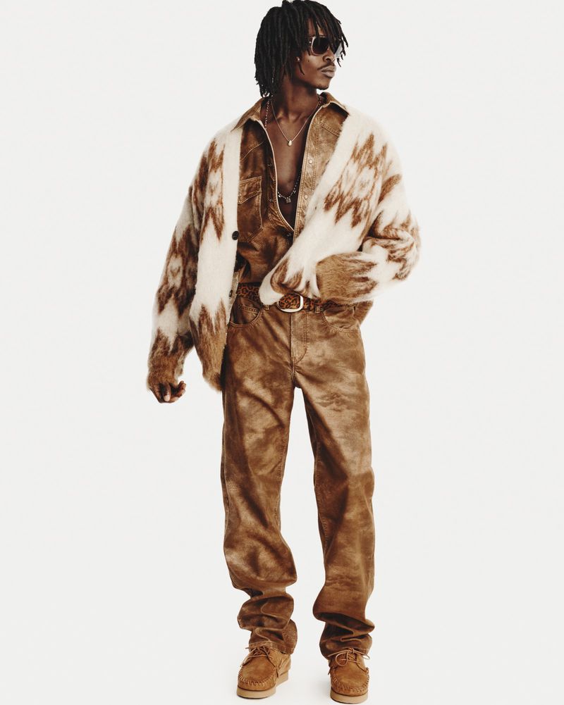 A black male model poses in a brown shirt, pants, cardigan, and sunglasses against a white backdrop in a fashion-forward studio shot.