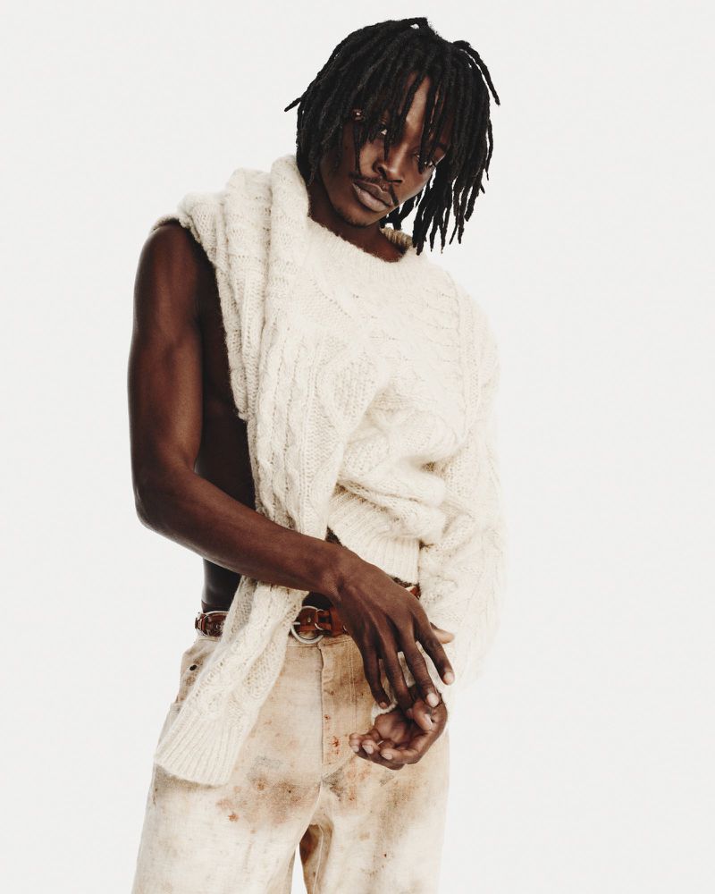 Man poses in a light off-shoulder sweater and stained pants against a white background. Fashion shot, eye-level angle, neutral expression.