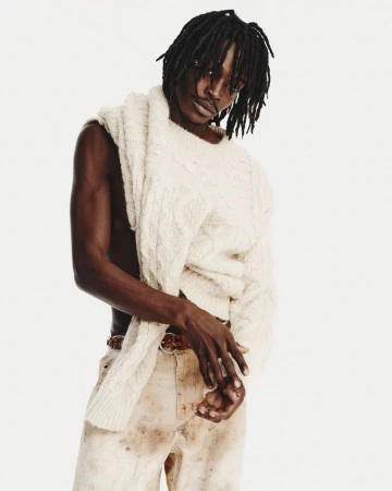 Man poses in a light off-shoulder sweater and stained pants against a white background. Fashion shot, eye-level angle, neutral expression.