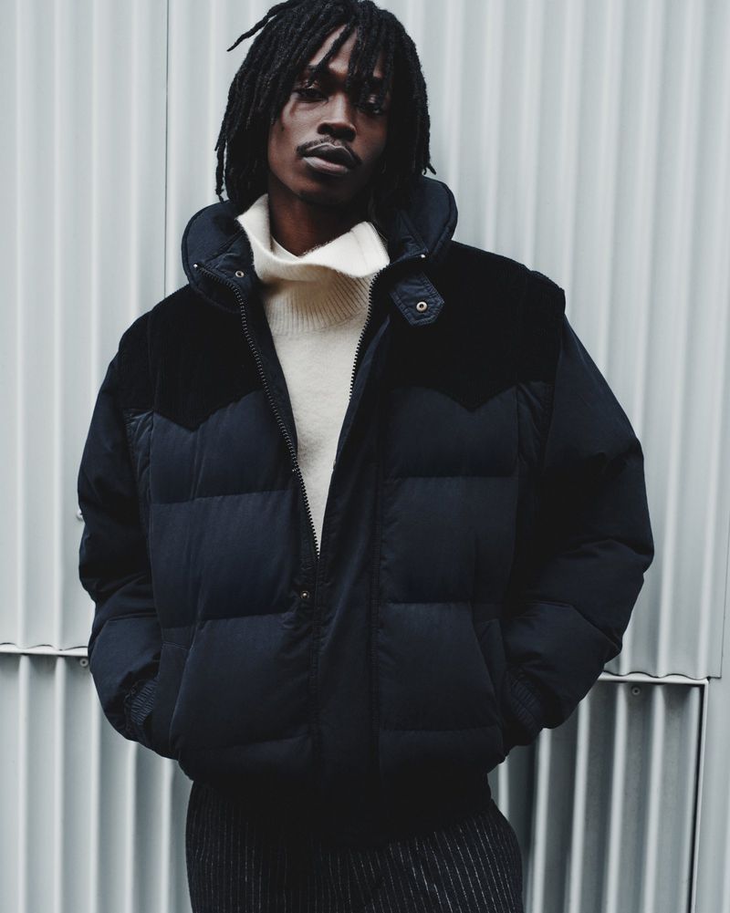 A single adult male model wears a navy puffer jacket and a cream turtleneck against a gray corrugated metal wall in a studio setting.