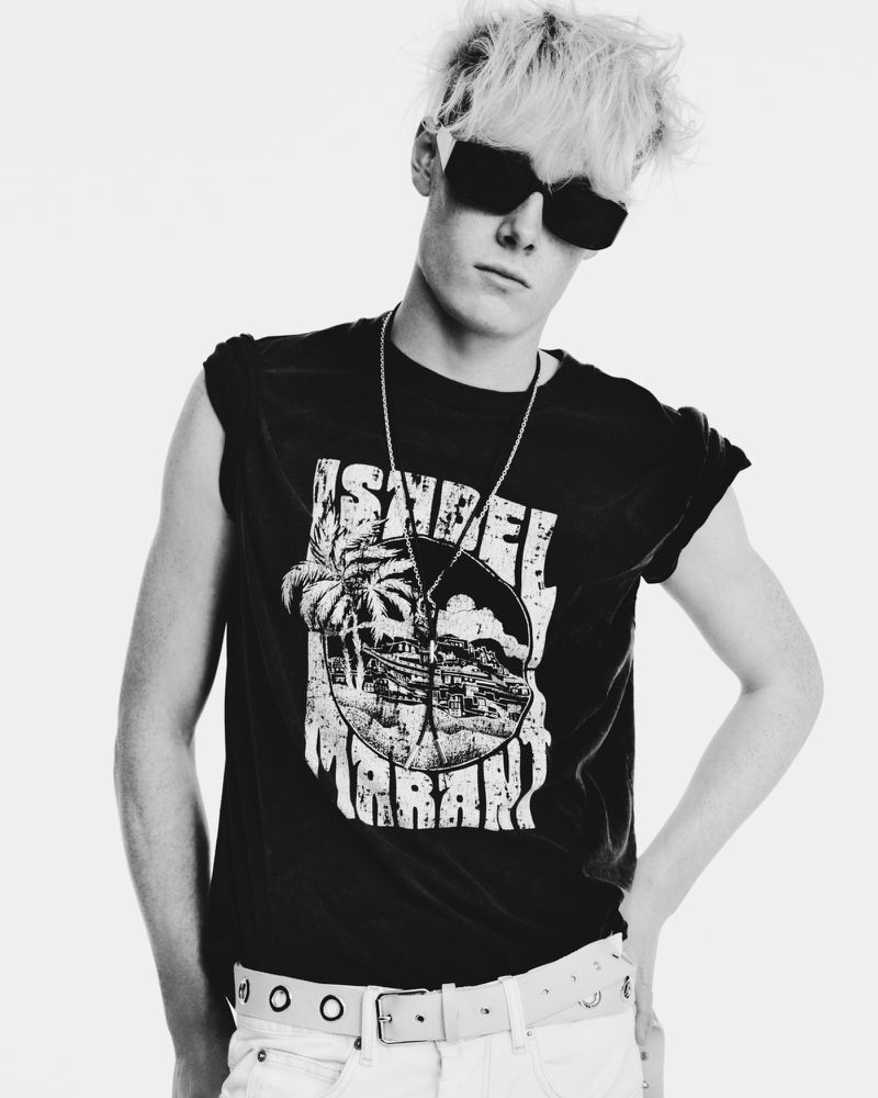 Monochrome portrait of a young adult male with blond hair wearing a graphic t-shirt, white jeans, and black sunglasses in a studio.
