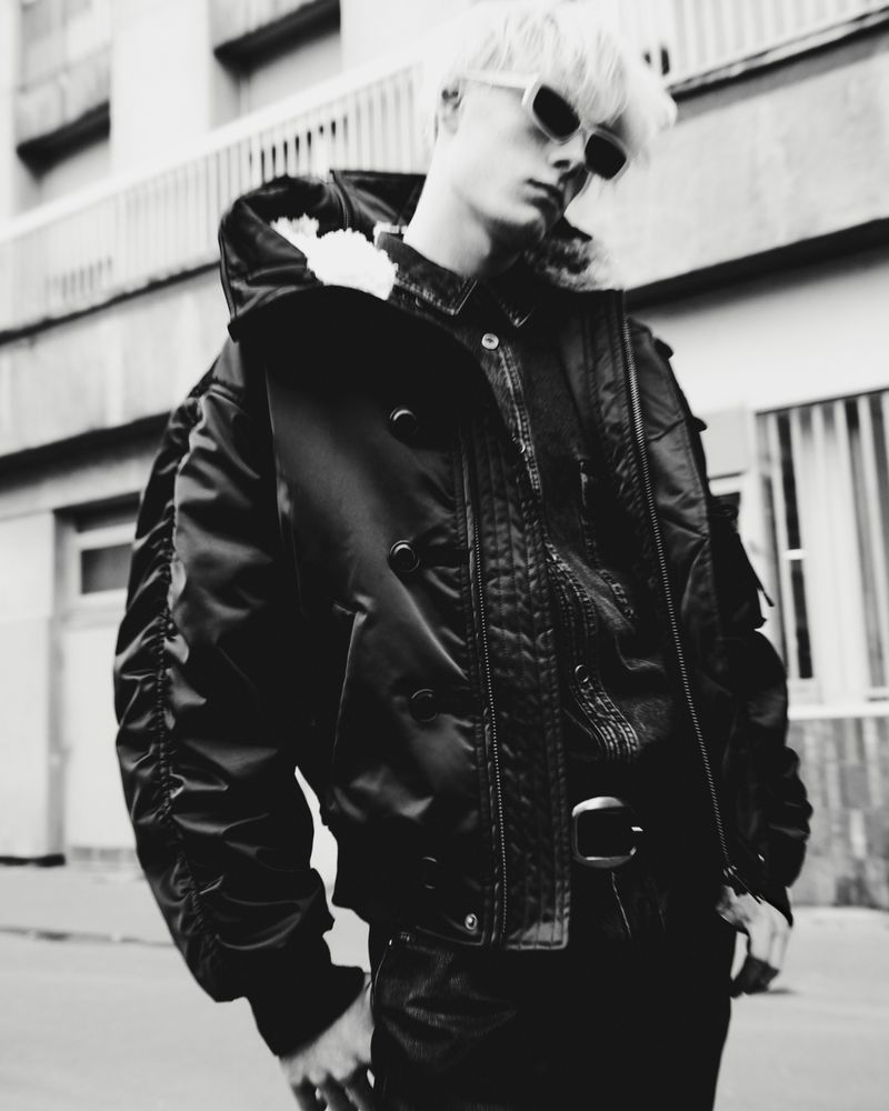 Monochrome portrait of a young adult with platinum hair, sunglasses, and bomber jacket standing outside a building in a city street.