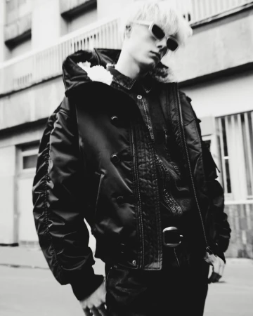 Monochrome portrait of a young adult with platinum hair, sunglasses, and bomber jacket standing outside a building in a city street.