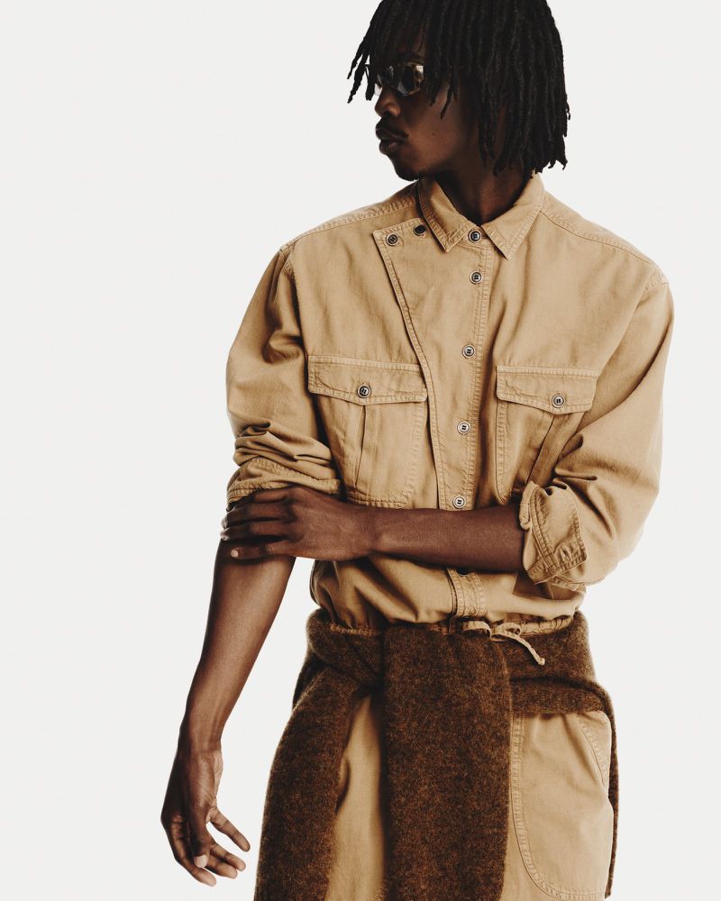 An adult Black male model poses indoors against a white background wearing a tan collared shirt and pants with a brown sweater tied around his waist.