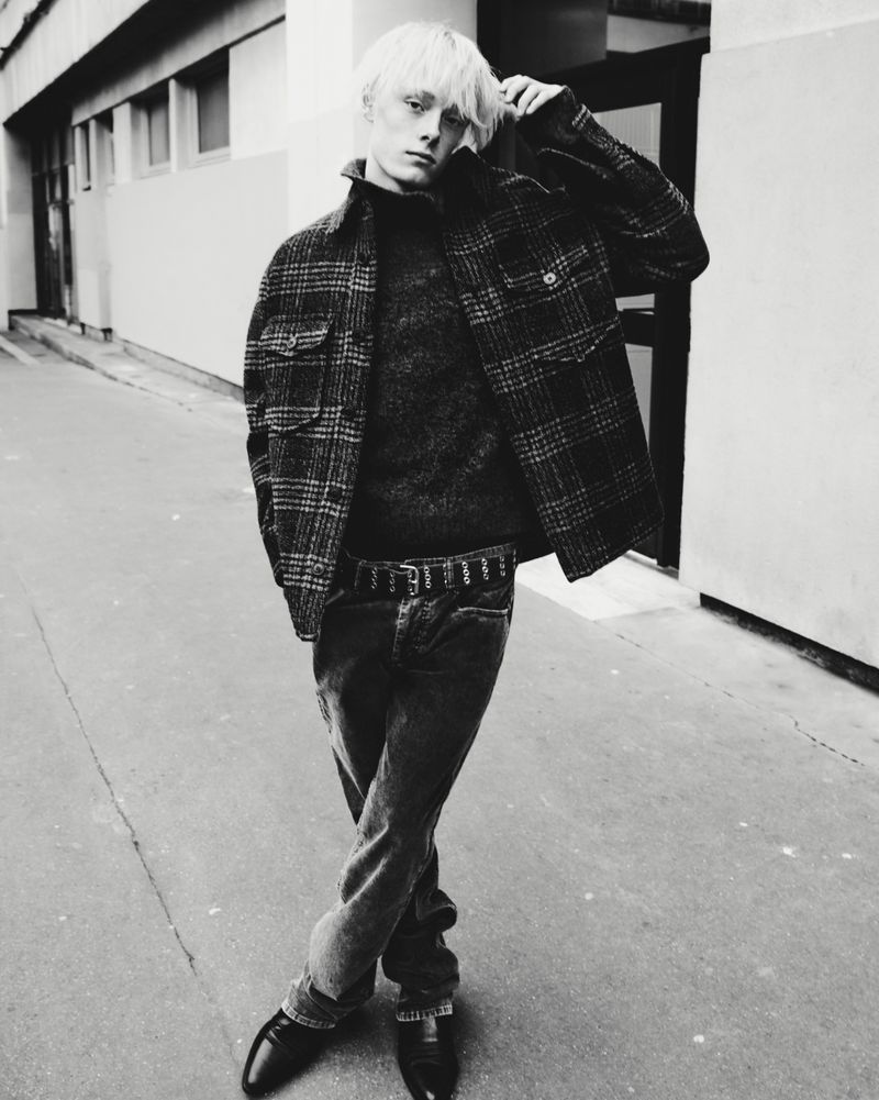 Black and white portrait of a male fashion model with fair hair wearing a wool jacket, sweater, jeans, and dress shoes outdoors.