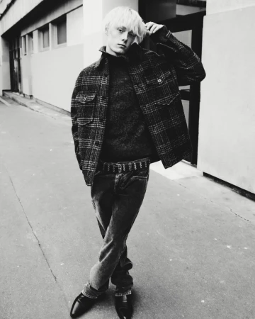Black and white portrait of a male fashion model with fair hair wearing a wool jacket, sweater, jeans, and dress shoes outdoors.