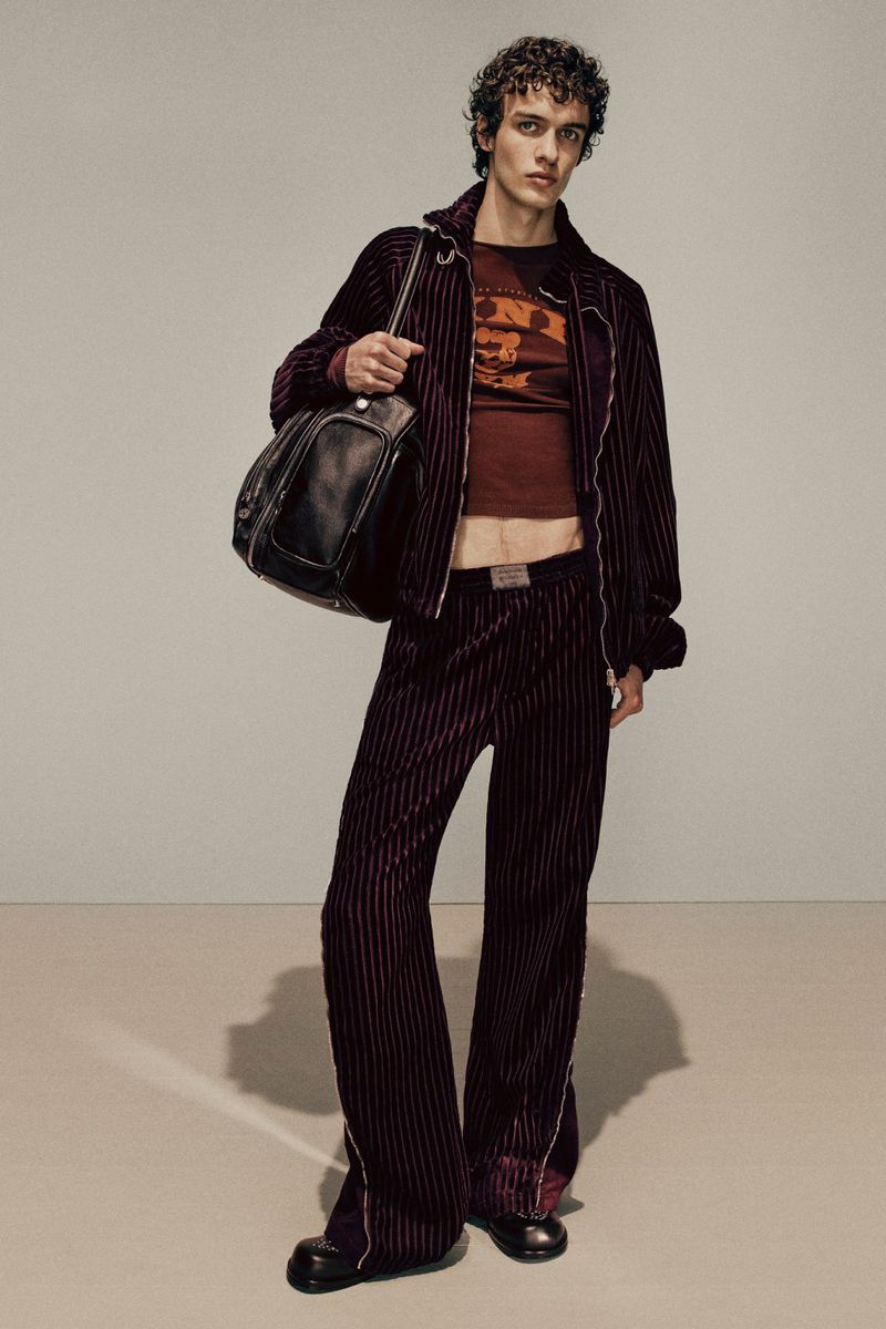 A slender male model in a maroon pinstripe velvet track suit, printed tee, and black leather duffel bag poses in a neutral studio setting.