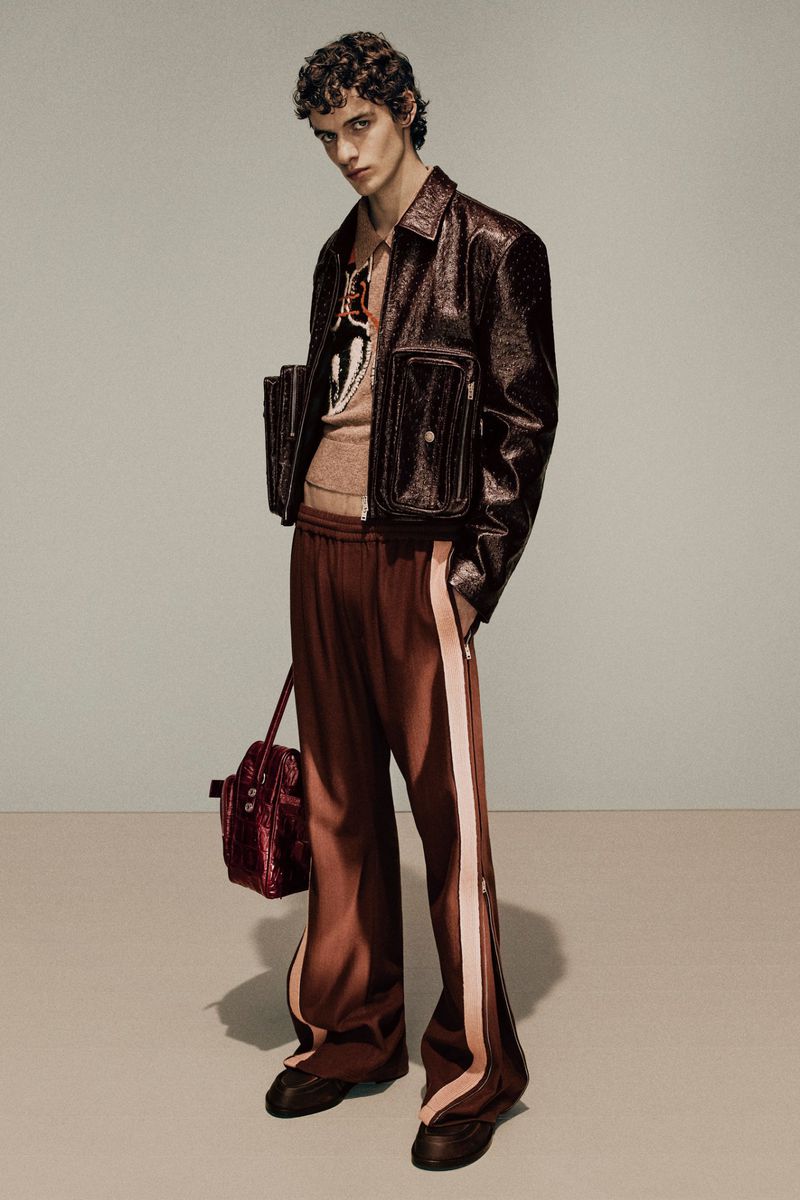 A young adult man poses in a studio wearing a brown jacket and pants while holding a matching shiny brown handbag.