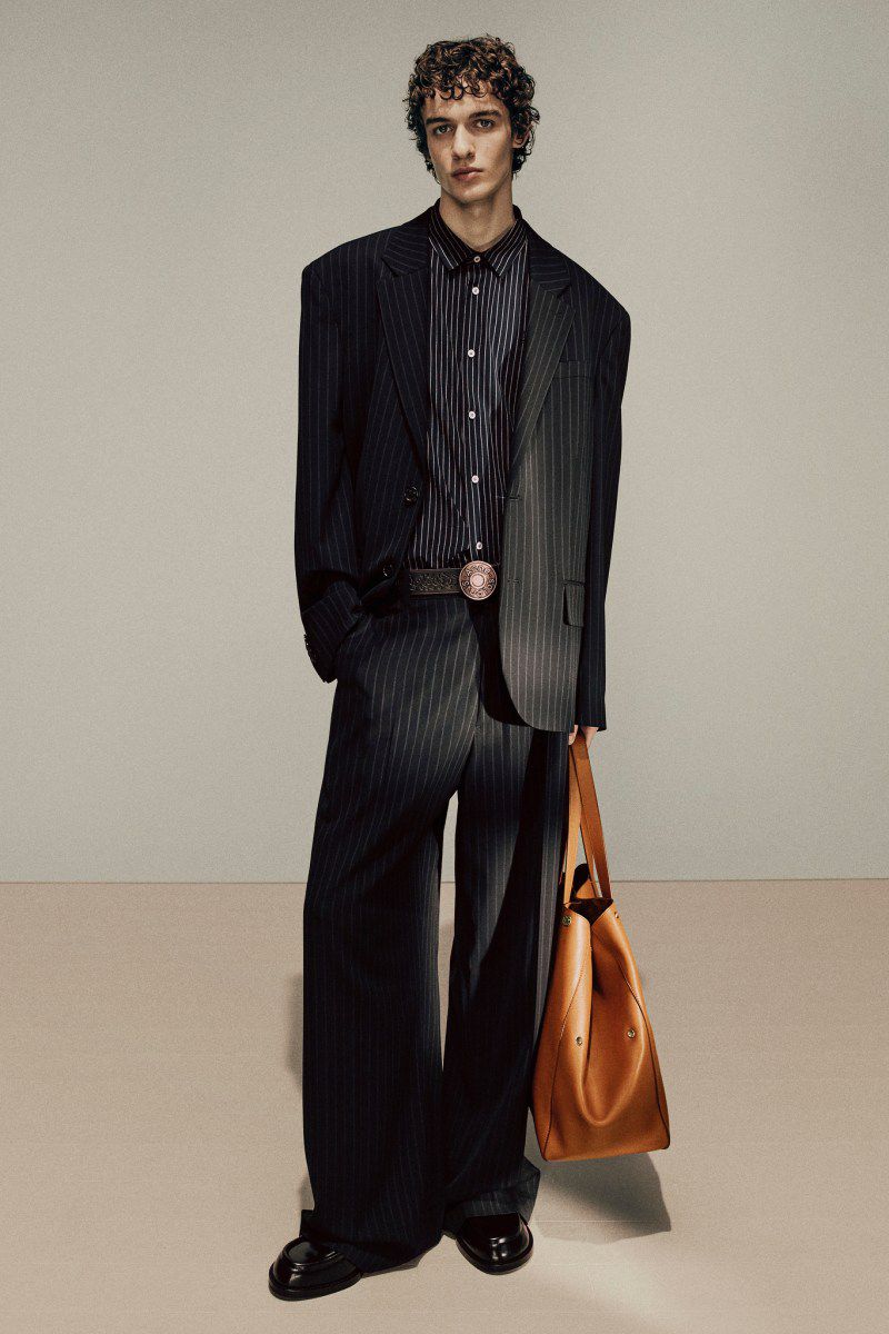 Male fashion model in pinstripe suit holding tote bag, posing in studio. Fashion editorial shot with neutral background and eye-level perspective.