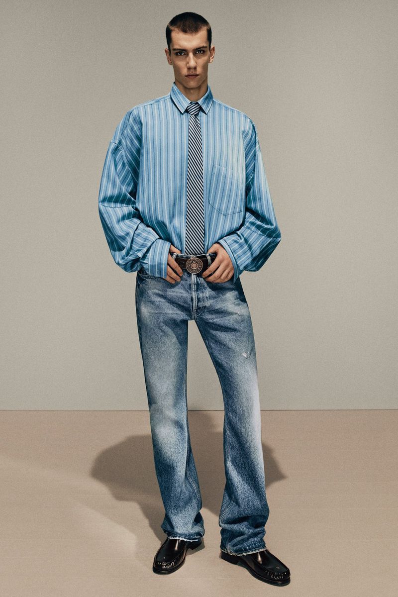 Fashion shot of a young adult male in a blue striped shirt, tie, jeans, and patent leather shoes, standing against a neutral backdrop.