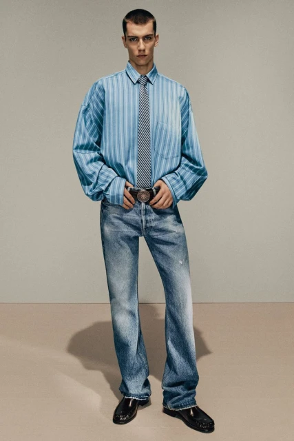 Fashion shot of a young adult male in a blue striped shirt, tie, jeans, and patent leather shoes, standing against a neutral backdrop.