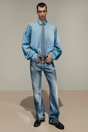 Fashion shot of a young adult male in a blue striped shirt, tie, jeans, and patent leather shoes, standing against a neutral backdrop.