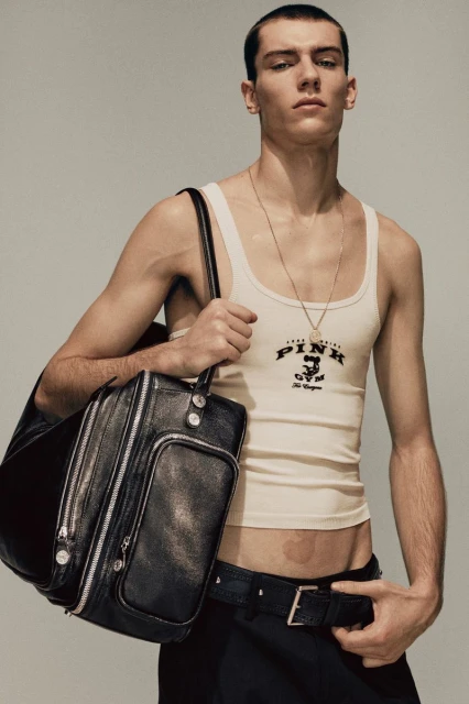 Portrait of a slender young man in a white tank top holding a black leather duffel bag against a light gray studio background. 