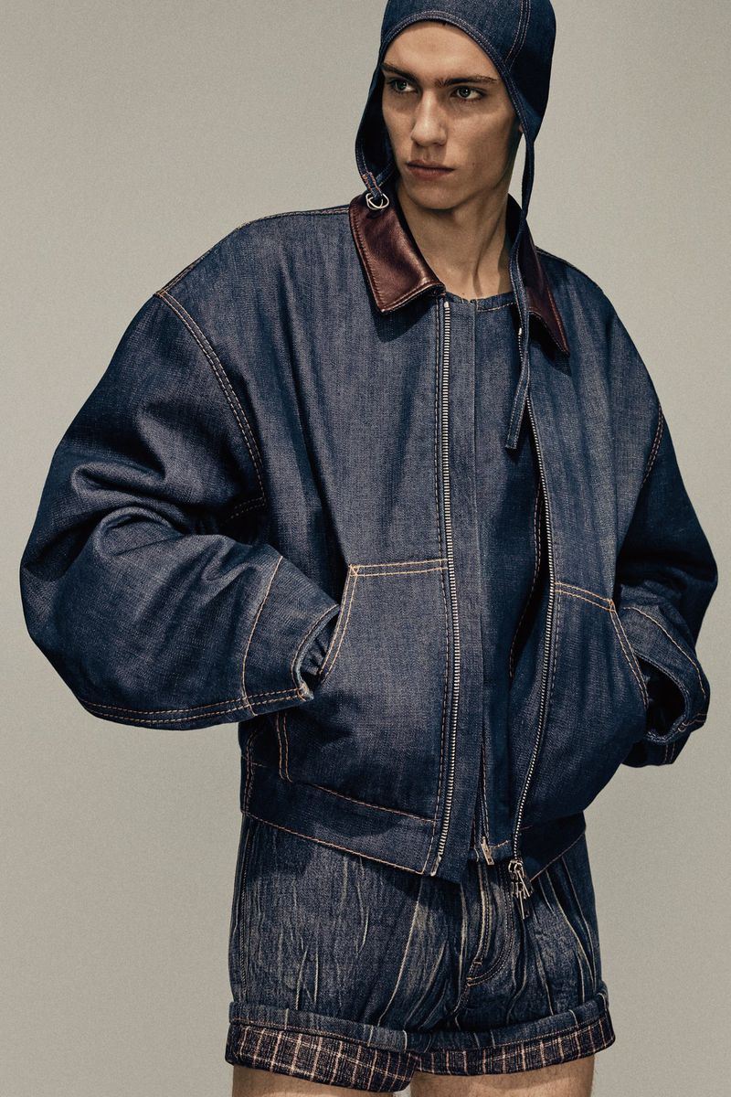 Fashion shot of a fair-skinned male model wearing a dark-wash denim jacket and shorts with a matching denim cap in a studio setting.