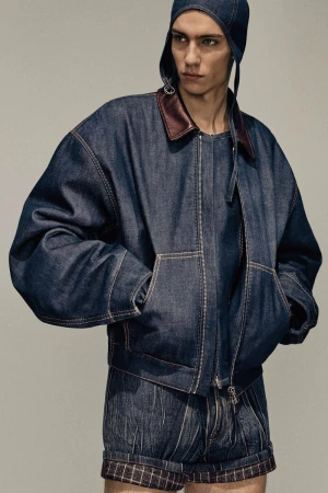 Fashion shot of a fair-skinned male model wearing a dark-wash denim jacket and shorts with a matching denim cap in a studio setting.